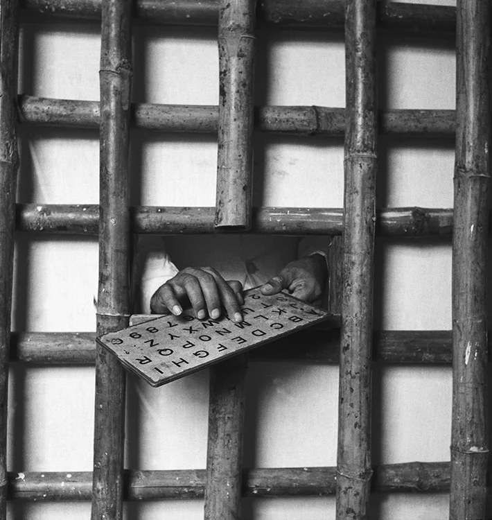 Meher Baba's hands passing an alphabet board through the bars of a bamboo screen
